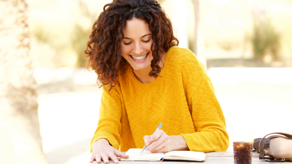 A brunette woman with curly hear wearing a yellow sweater writing on a notebook outdoors